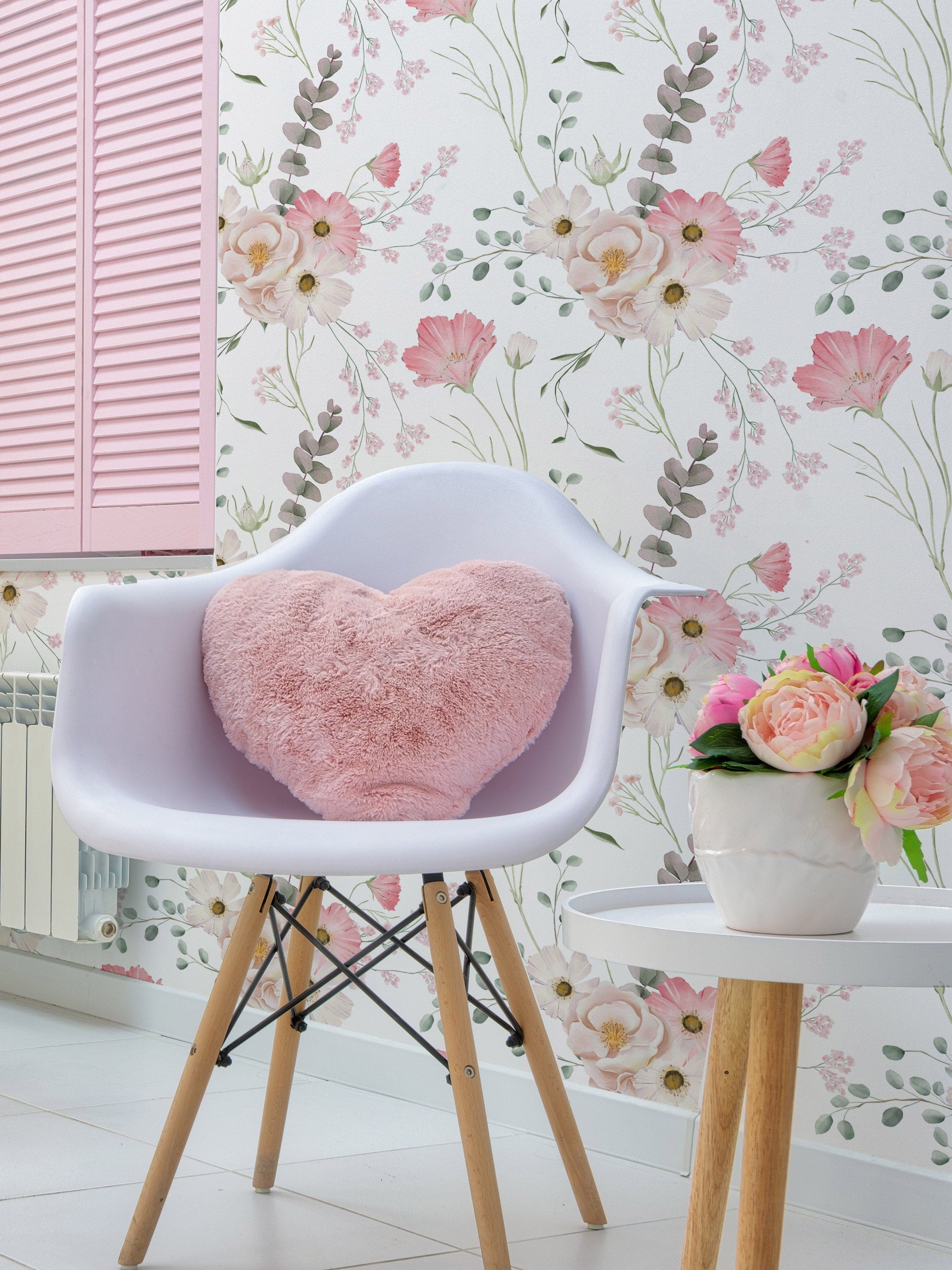 Flourish Wallpaper in a cozy living space featuring a white chair with wooden legs, a pink heart-shaped cushion, a side table with a vase of pink flowers, and a soft floral wallpaper with delicate pink and white flowers and green foliage