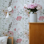 Flourish Wallpaper in a charming bedroom setting with a wooden dresser, a white wall sconce, and a vase of pink flowers. The wallpaper features a soft floral design with pink and white flowers and green foliage.