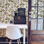 A stylish home office featuring a white desk and chair, set against a wall adorned with Vintage Branches Wallpaper. The wallpaper's intricate design of green leaves and brown branches on a white background adds a natural and refreshing touch to the workspace.