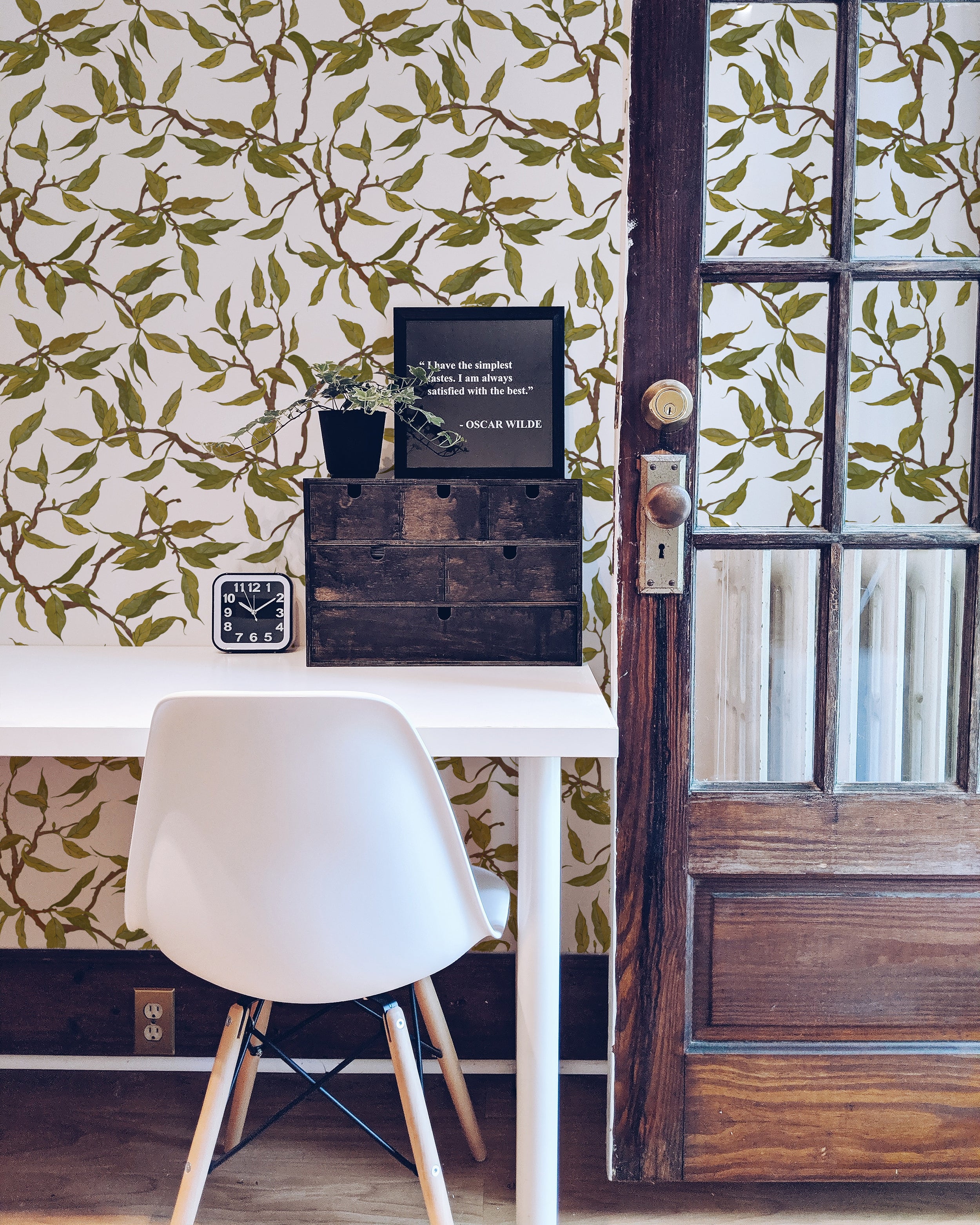 A stylish home office featuring a white desk and chair, set against a wall adorned with Vintage Branches Wallpaper. The wallpaper's intricate design of green leaves and brown branches on a white background adds a natural and refreshing touch to the workspace.