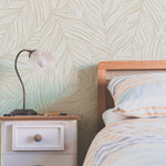 Interior bedroom scene with a bed partially visible on the right, dressed with striped linens in muted orange and blue. A small, vintage-style white nightstand with a single knobbed drawer sits beside it, supporting a curved metal lamp with a tulip-shaped white shade. The wall behind features a large-scale wallpaper with an abstract leaf pattern in soft ecru tones, adding a botanical and tranquil feel to the space.