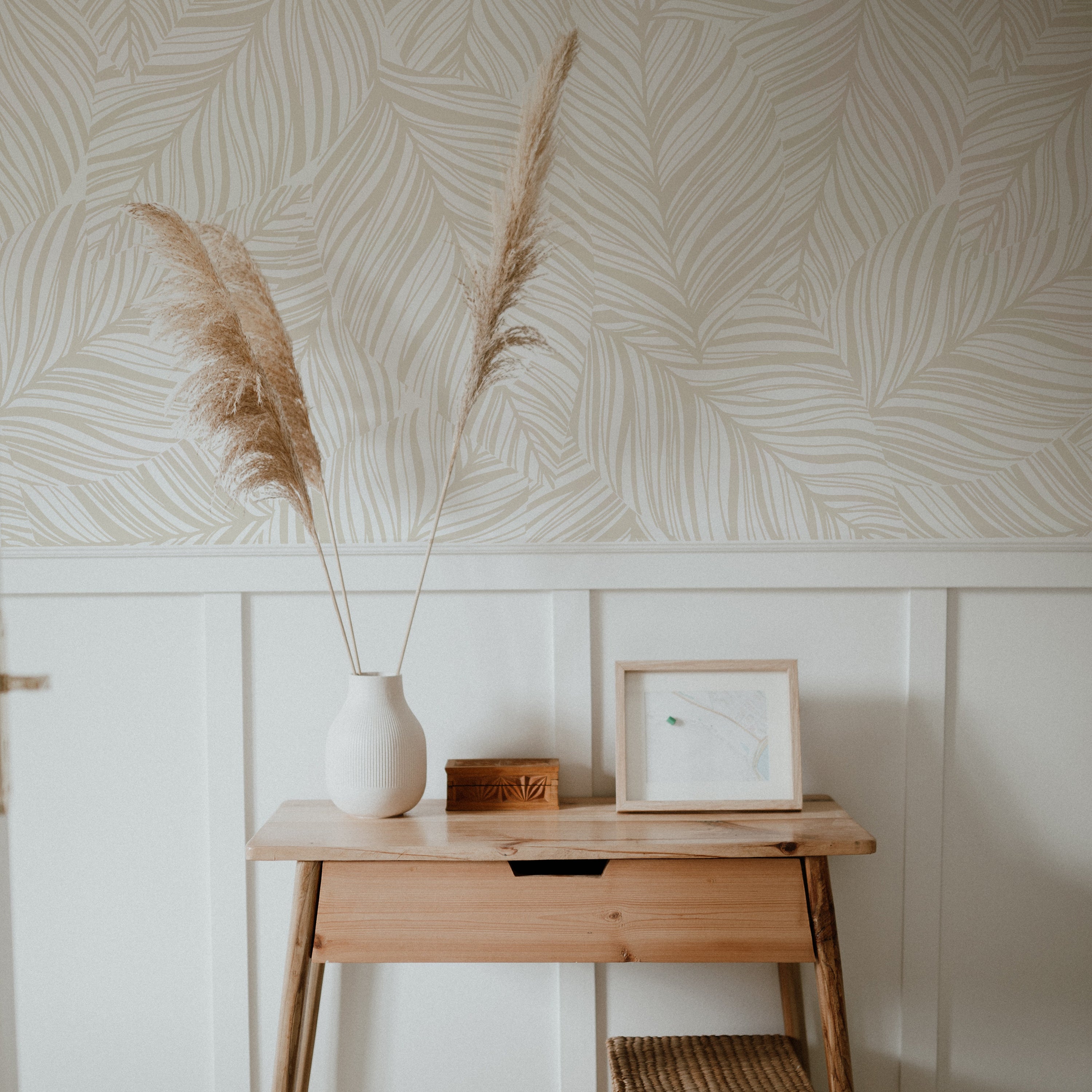 A cozy corner of a room featuring the same ecru leaf-patterned wallpaper as a backdrop. In the foreground, a simple wooden console table holds a tall, slender white vase with two pampas grass stems, complementing the wallpaper's organic theme. A framed abstract artwork and a small wooden box rest beside the vase, against a white wainscoting that adds architectural detail.