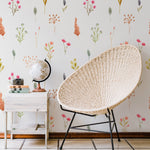 A cozy corner featuring the Watercolour Floral Wallpaper II, adorned with vibrant watercolor flowers in pinks, oranges, and greens on a cream background. A wicker chair and a small wooden table with a vintage globe and camera add a touch of nostalgia to the whimsical setting.