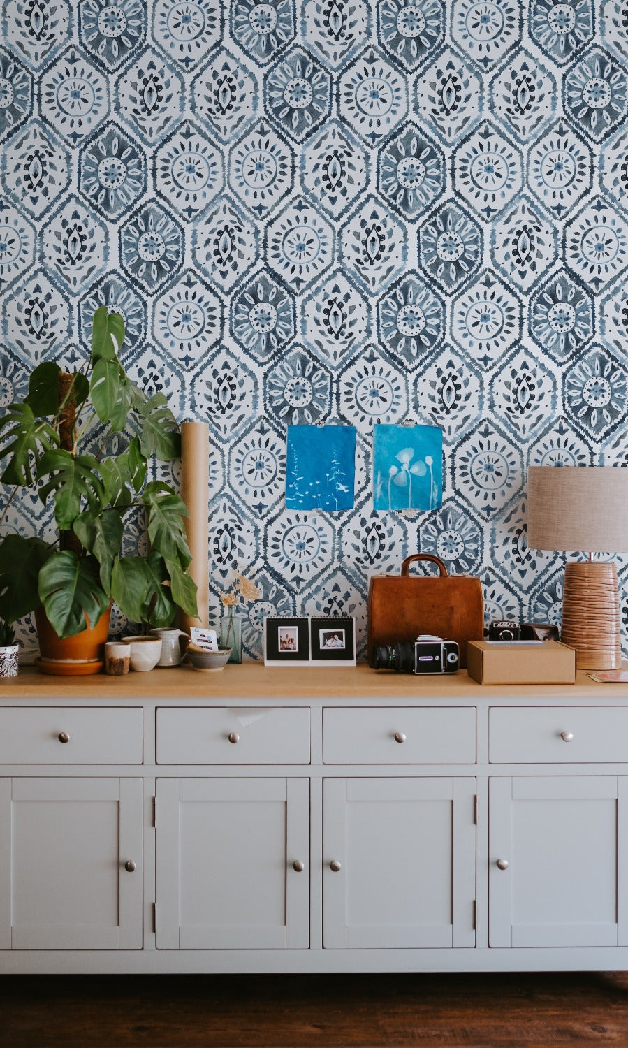 A stylish interior design featuring a white cabinet with drawers and doors against a Moroccan Tile wallpaper in indigo blue, decorated with plants, framed photos, and various knick-knacks.