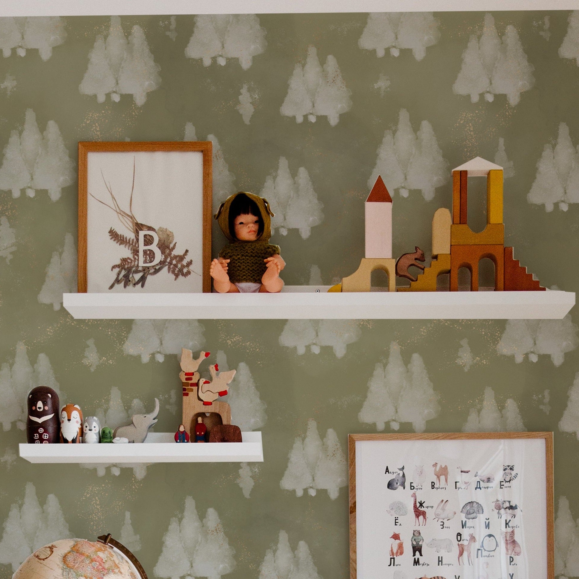 A stylish room featuring Shimmer Tree Wallpaper, showcasing a pattern of white snowy trees on a muted olive green background, with sparkling gold accents giving a festive and magical look. The room is decorated with toys and educational items on white shelves, enhancing the cozy, wintery theme.
