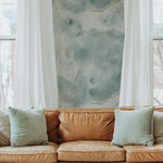 Cozy living room setup featuring a Shimmering Marble Wallpaper with a unique blue and green watercolor pattern and elegant gold veins. The wallpaper is complemented by a tan leather sofa and soft blue pillows, framed by white drapes