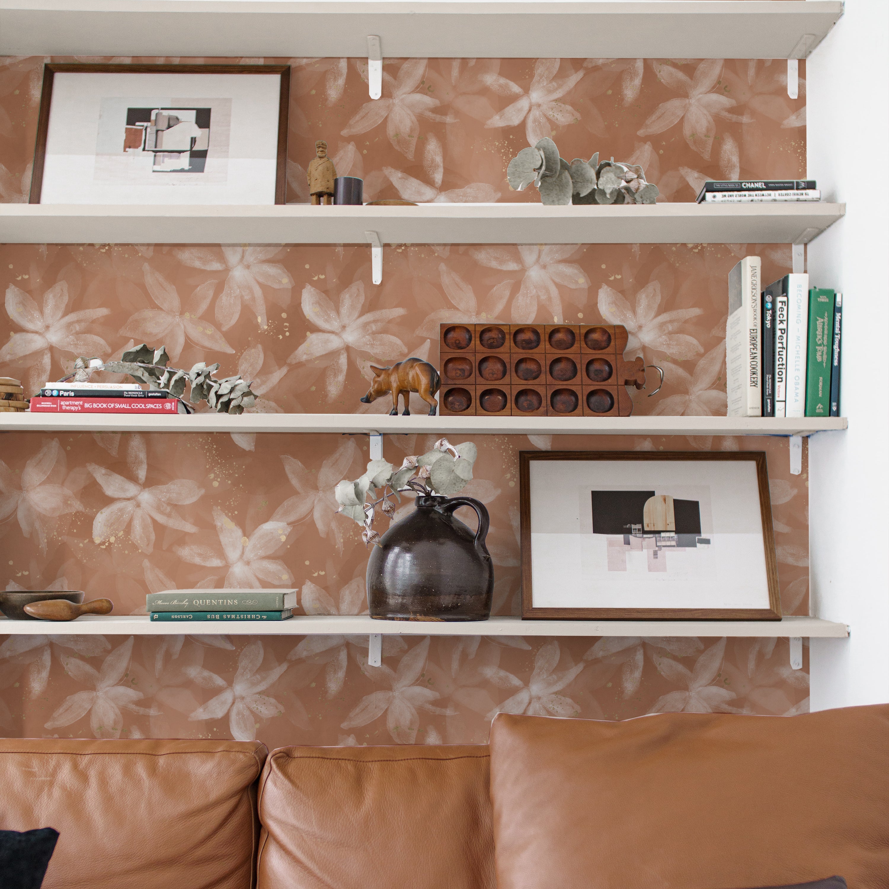 A stylish living room with Shimmer and Terracotta Wallpaper accenting the wall behind white shelves. The shelves are decorated with books, sculptures, and pottery, which complement the earthy tones of the wallpaper. The pattern of translucent flowers adds a delicate touch to the room's decor