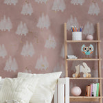 A child's bedroom displaying the Shimmer Tree Wallpaper - Pink on the walls, which portrays whimsical white trees with a snowy texture against a pastel pink backdrop, complemented by gold specks. The room is styled with cozy bedding and a playful assortment of toys and books, enhancing the room's gentle and magical atmosphere