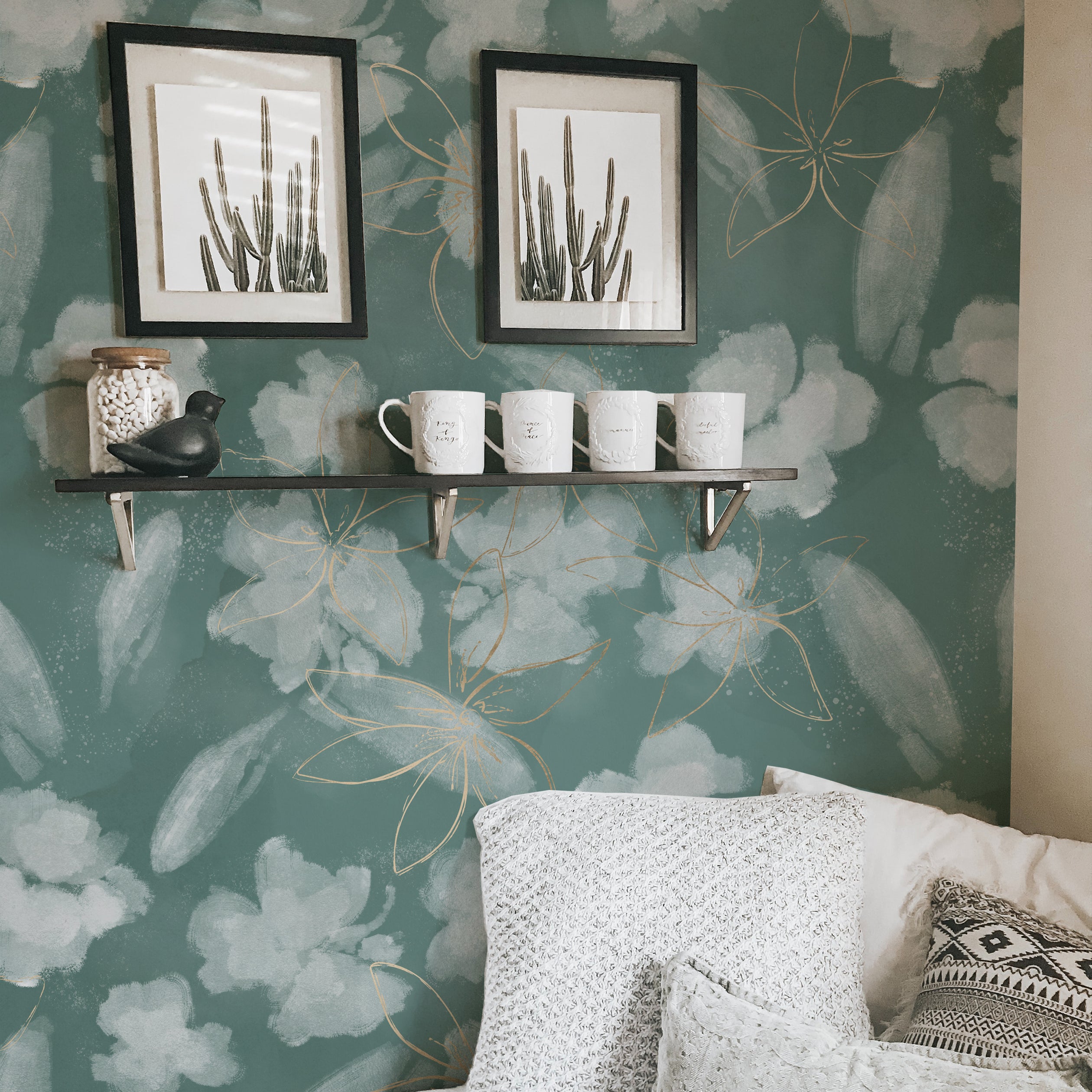 A cozy corner of a home decorated with Shimmer and Gold Wallpaper, enhancing the wall behind a floating shelf displaying cactus prints, assorted mugs, and a small decorative bird. The wallpaper's green backdrop and gold accents complement the earthy and neutral colors of the decor.