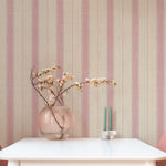 A dining room featuring walls adorned with vertical-striped wallpaper in beige and muted pink. The room includes a white dining table, pink chairs, and a vase with flowers, creating a warm and inviting ambiance.