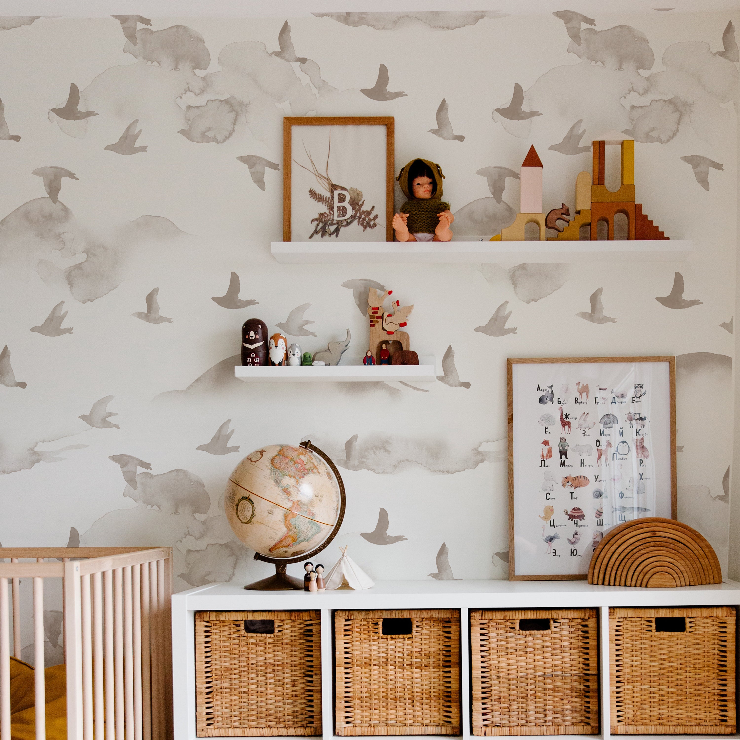 a cozy children's room decorated with the Kids Wild Pinery Wallpaper. The wallpaper's bird motifs provide a soothing, nature-inspired backdrop that complements the room's warm wooden furniture, including floating shelves adorned with toys and educational frames. The room is grounded by wicker storage baskets and a classic globe, fostering a nurturing environment for growth and learning.