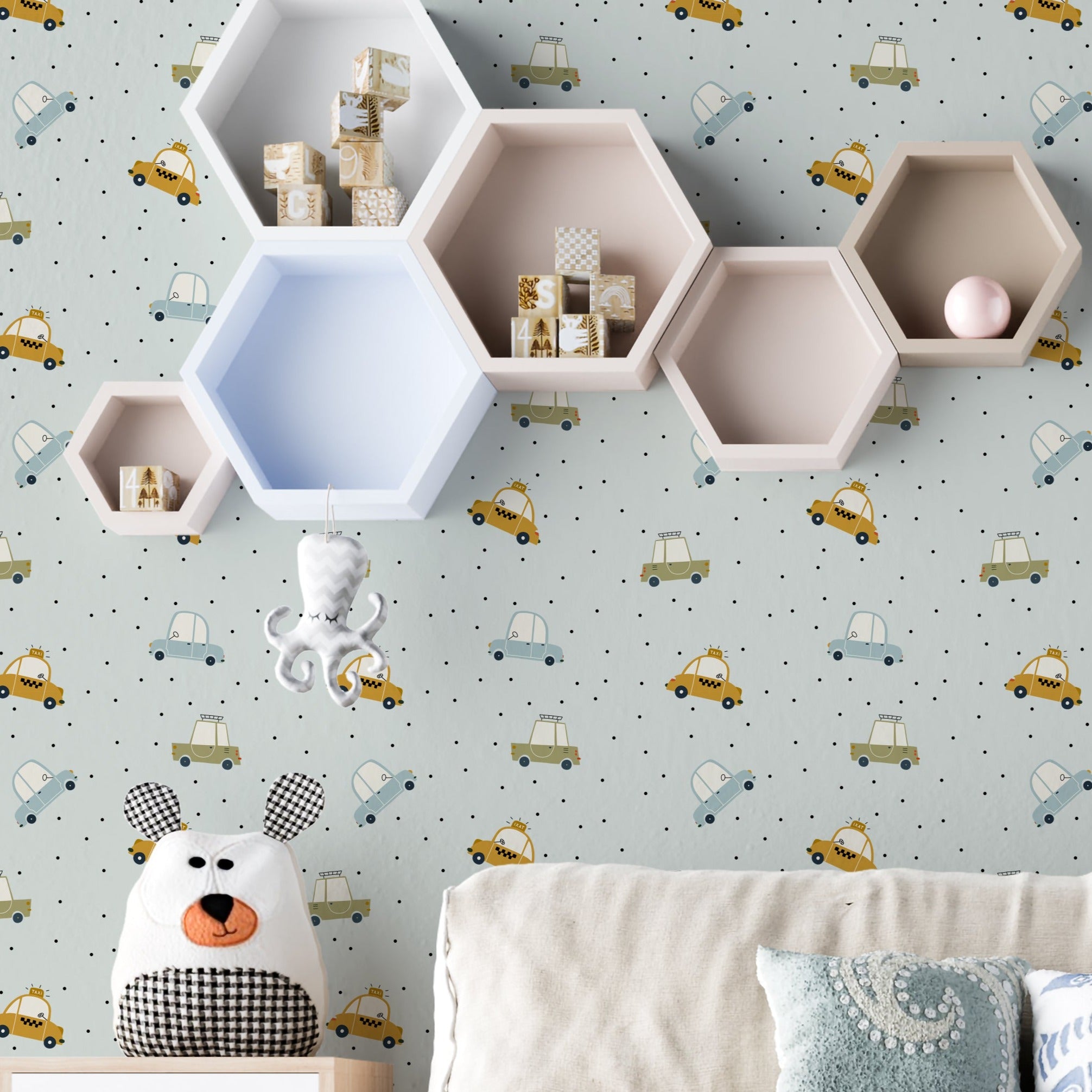 A cozy children's room setting showcasing the Cute Cars Wallpaper, with cheerful blue and yellow cars scattered across a dotted gray backdrop. The playful theme complements the room's decor, including a stuffed bear, wooden toys, and a modern, child-friendly furniture setup