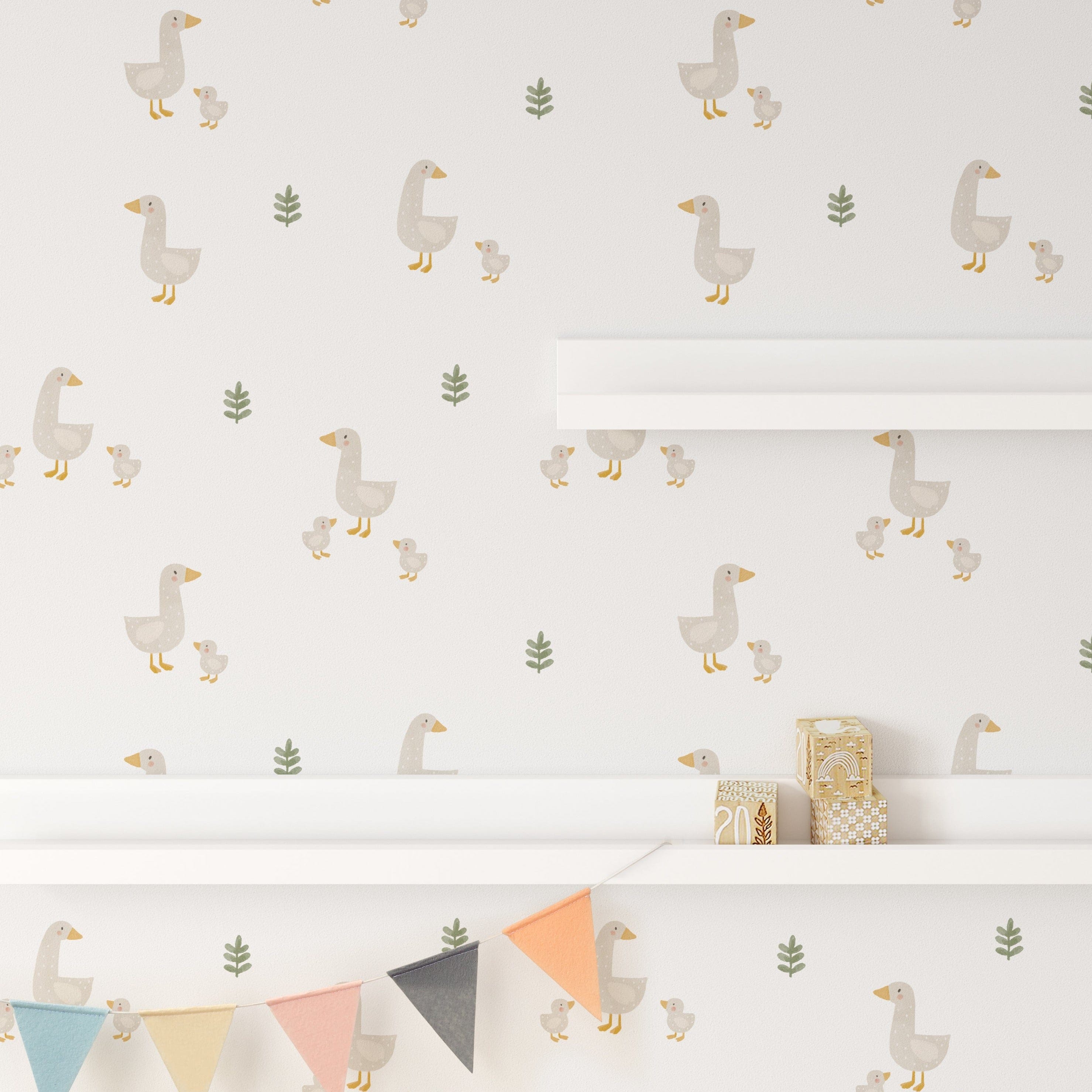 A section of a nursery wall covered in the Cute Farm Friend Wallpaper with a pattern of ducks and green plants. The wallpaper is accentuated by a colorful bunting, a white shelf with children's toys and books, offering a playful yet serene backdrop for a child's room.