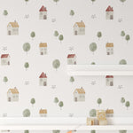 A section of a wall decorated with Farm Friend Wallpaper - Happy House, featuring charming houses and trees, along with a white shelf and colorful pennant banner, adding a cheerful touch to a child's room.