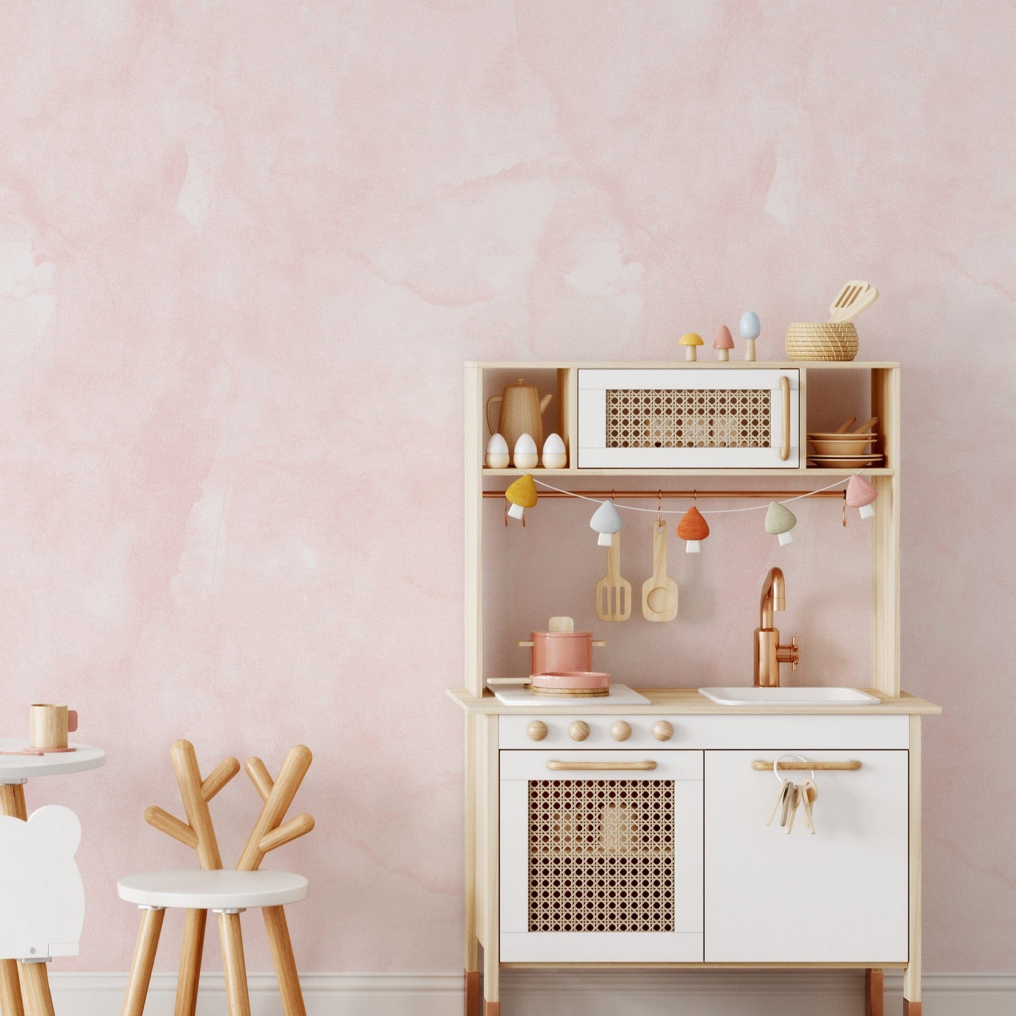 Children's play kitchen set against a wall covered in Kids Lime Wash Wallpaper - Blush Pink. The wallpaper's soft watercolor texture complements the wooden kitchen toys, creating a warm and playful environment
