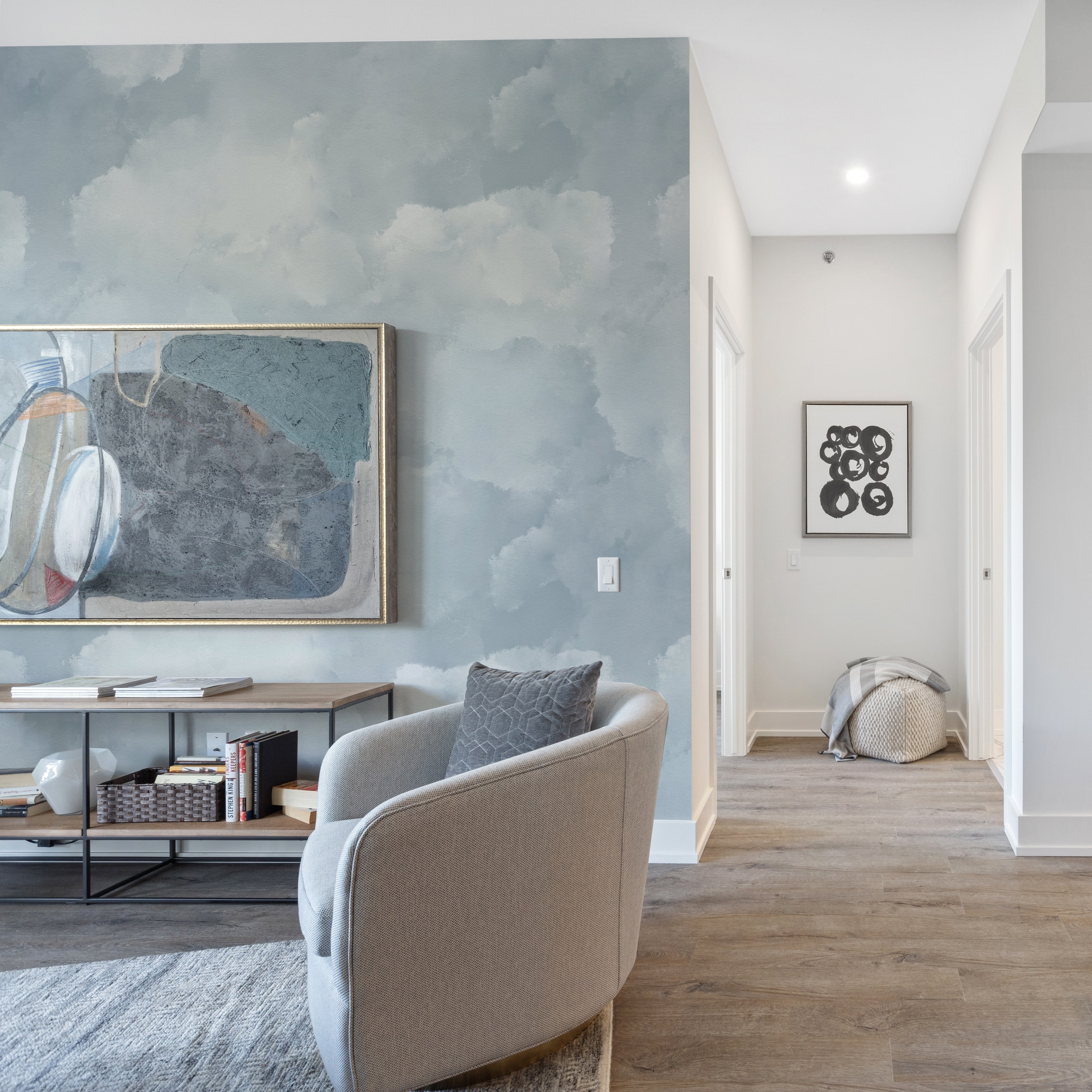 A modern living space featuring the Cloud Mural Wallpaper in Pale Blue on the wall, complementing a contemporary art piece and stylish furnishings. The wallpaper's soft cloud pattern enhances the room's airy and open feel