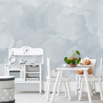 A children's play area with Cloud Mural Wallpaper in Pale Blue adorning the walls, evoking a dreamy sky with soft, fluffy cloud patterns. The serene pale blue tones offer a calming backdrop to the crisp white children's furniture in the room