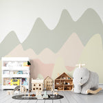 Pastel Hills Mural in a children's playroom featuring wavy pastel hill designs in soft shades of green, pink, and beige. The room is decorated with a plush elephant rocker, wooden toy houses, and a white bookshelf filled with children's books.
