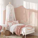 Pretty Hills Mural in a cozy kids' bedroom with a white bed draped with a canopy, pink and white bedding, a wicker basket, and a white desk. The wallpaper showcases soothing wavy pastel hills in shades of pink and beige.