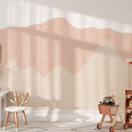 Pretty Hills Mural in a children's playroom featuring wavy pastel hill designs in soft shades of pink and beige. The room is decorated with a white play table and chair, a toy market stand, and a wooden toy cart filled with flowers.