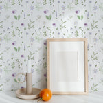 A serene space decorated with the Wildflower Wonder Wallpaper, featuring its delicate wildflower pattern in purple and yellow. The decor includes a framed picture, a candle, and a fresh orange on a white table