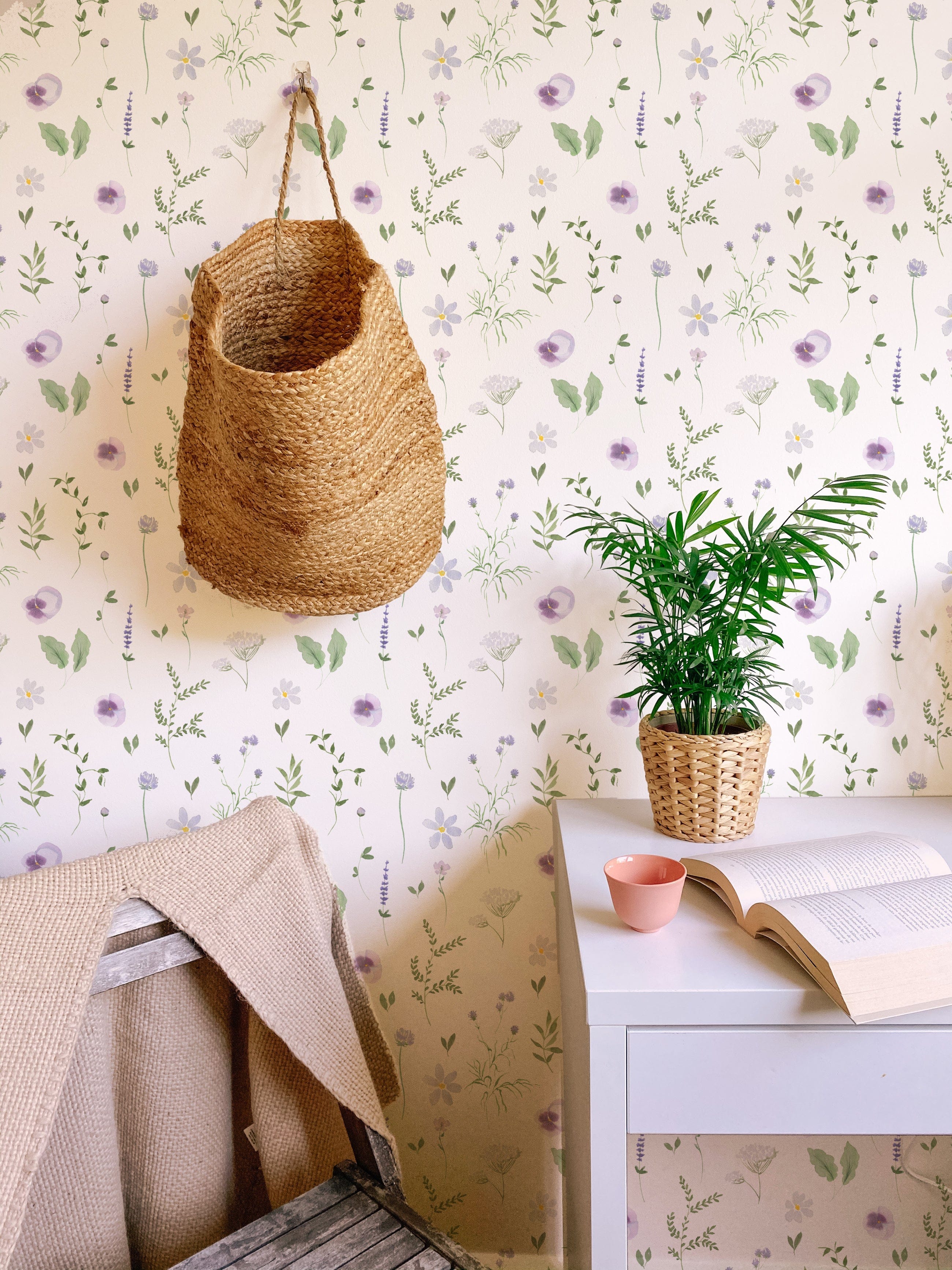A cozy room featuring the Wildflower Wonder Wallpaper with a soft white background adorned with delicate purple and yellow wildflowers and green foliage, complemented by a woven basket hanging on the wall and a small potted plant on a white desk.