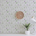 A workspace decorated with the Wildflower Wonder Wallpaper, showcasing its charming pattern of purple and yellow wildflowers and green leaves. The space includes a wooden wall clock and a white desk lamp beside a small plant.