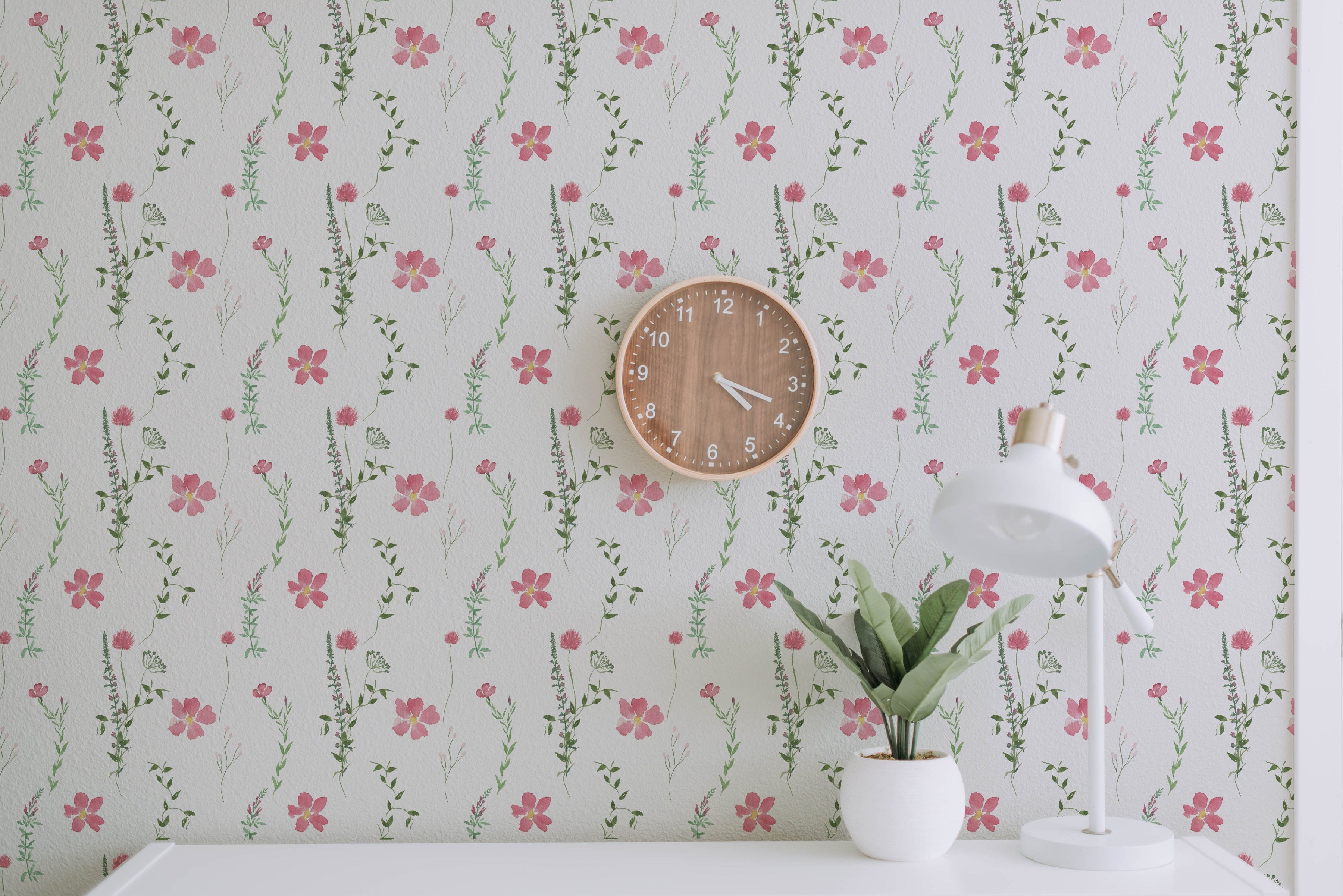 A room with a Spring Serenity Wallpaper featuring a white background adorned with delicate pink flowers and green leaves, complemented by a wooden wall clock and a white desk lamp on a desk with a small potted plant