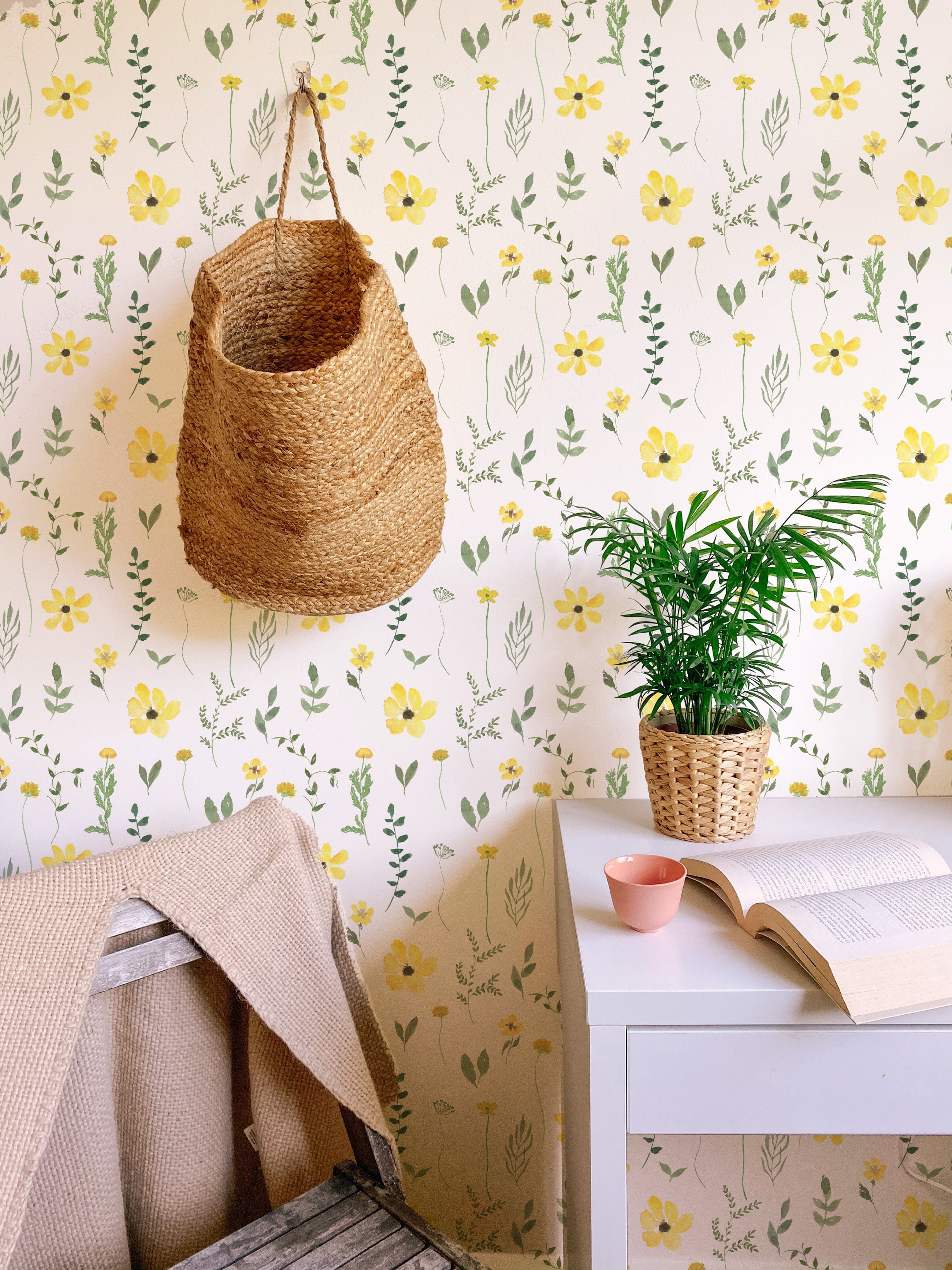 A cozy room featuring the Spring Field Wallpaper - V with a soft white background adorned with bright yellow flowers and green leaves, complemented by a woven basket hanging on the wall and a small potted plant on a white desk