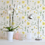 A stylish room corner with the Spring Field Wallpaper - V, highlighting its cheerful yellow flower pattern. The decor includes a white orchid in a pot and a sleek metallic desk lamp on a white desk