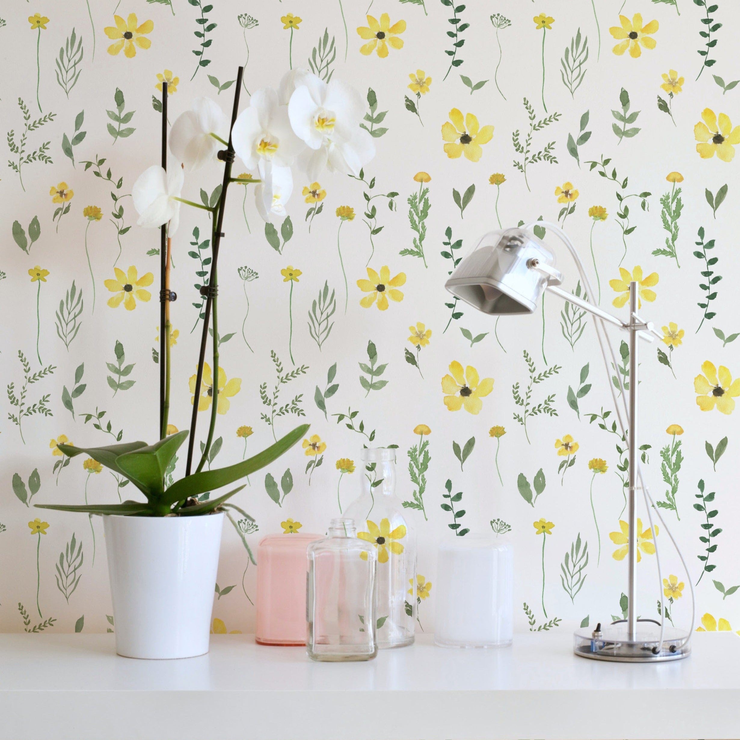 A stylish room corner with the Spring Field Wallpaper - V, highlighting its cheerful yellow flower pattern. The decor includes a white orchid in a pot and a sleek metallic desk lamp on a white desk