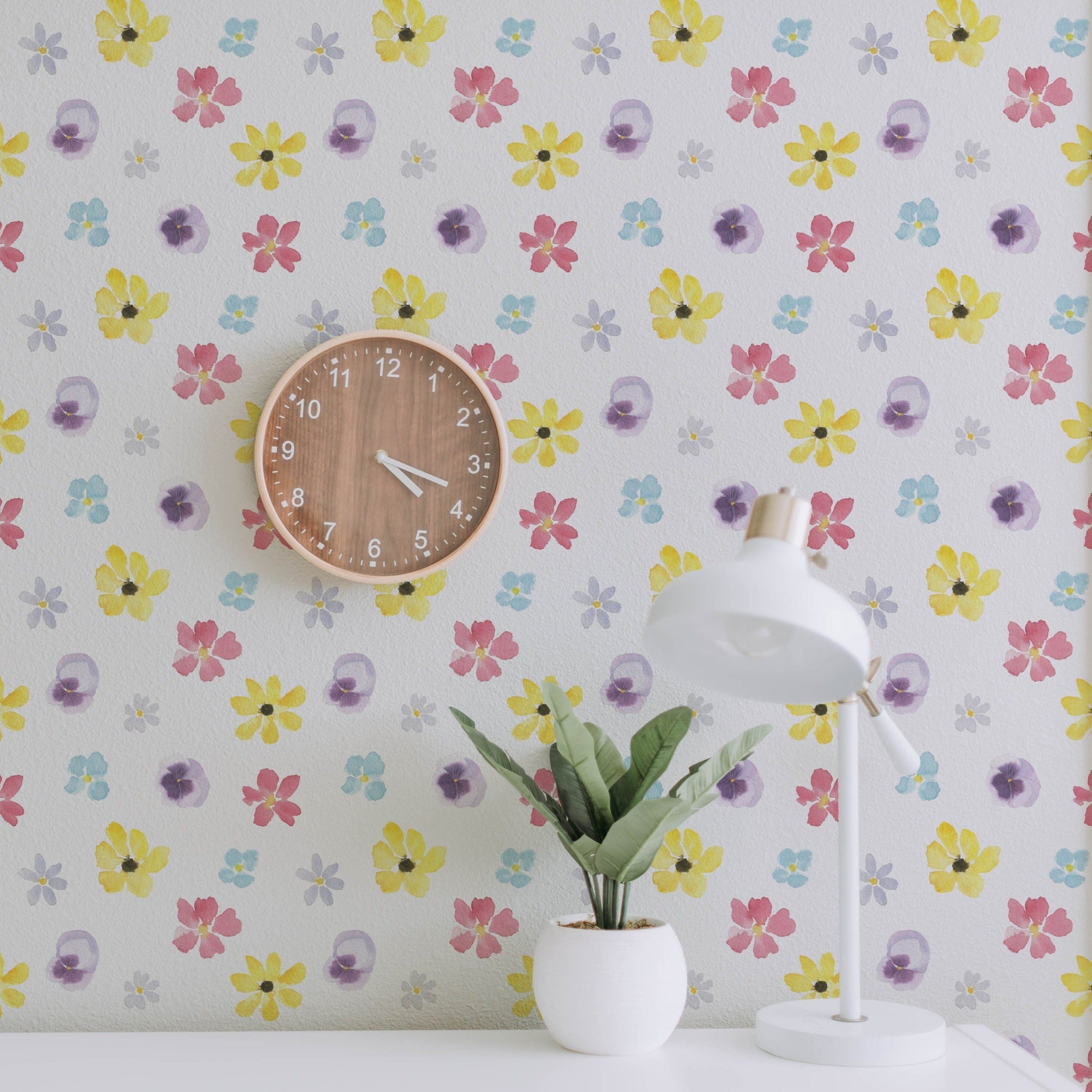 A workspace adorned with the Spring Field Wallpaper - VI, highlighting its lively floral pattern in bright yellow, pink, purple, and blue hues. The space includes a wooden wall clock and a white desk lamp beside a small plant.