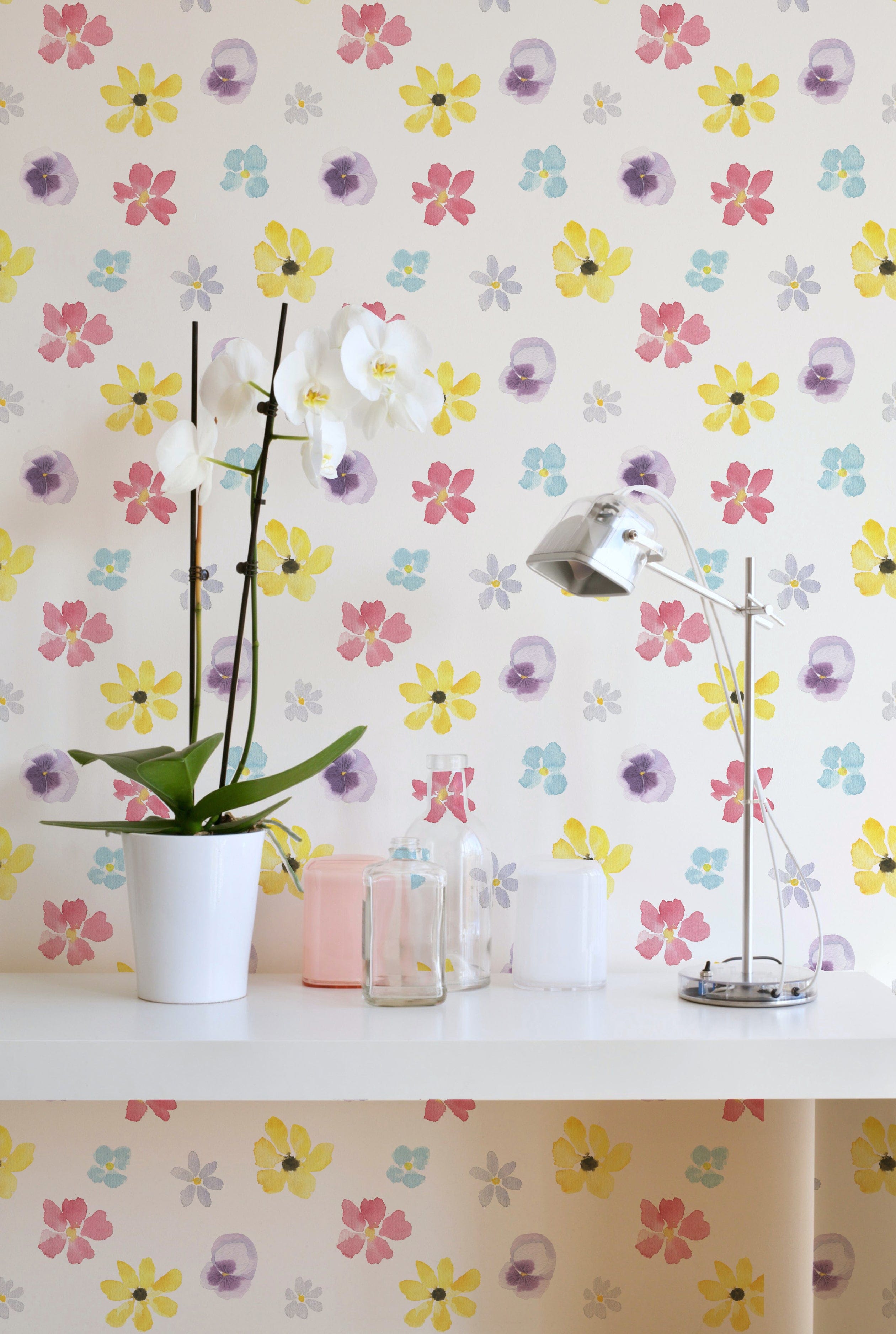 A cozy room featuring the Spring Field Wallpaper - VI with a cheerful pattern of colorful watercolor-style flowers in yellow, pink, purple, and blue on a white background. The decor includes a white orchid in a pot and a sleek metallic desk lamp on a white desk.