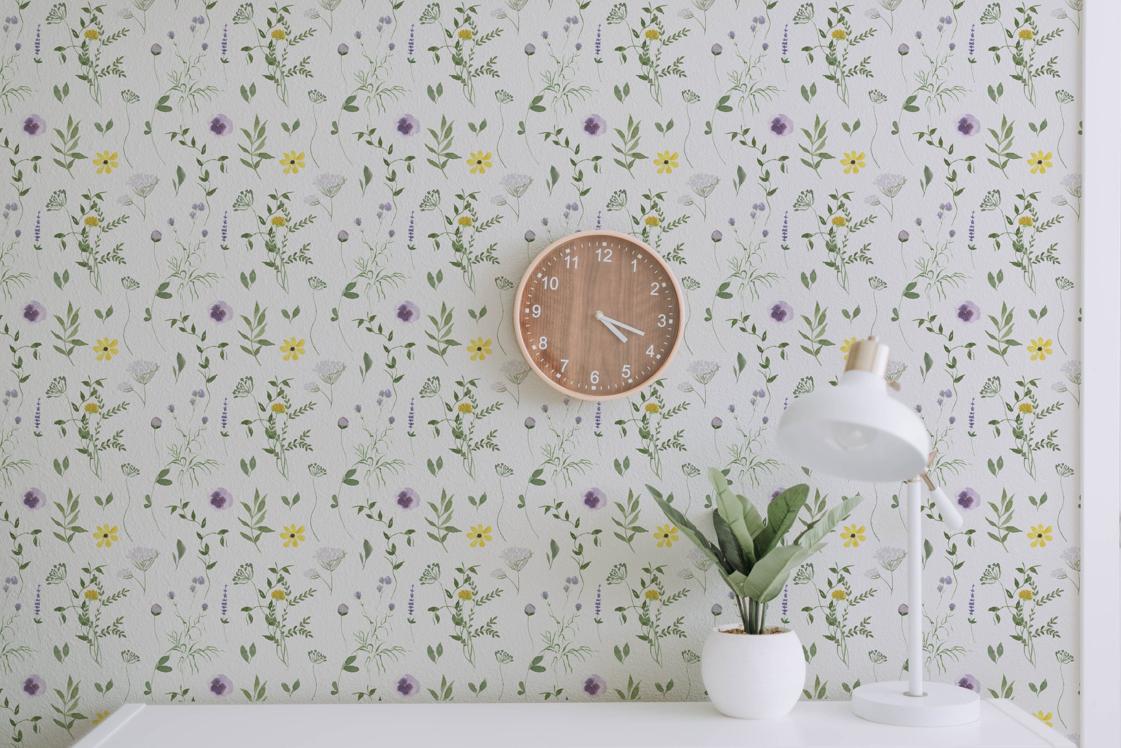 A workspace featuring the Spring Field Wallpaper - VII with a white background adorned with delicate purple and yellow flowers and green leaves. The decor includes a wooden wall clock and a white desk lamp beside a small plant in a white pot
