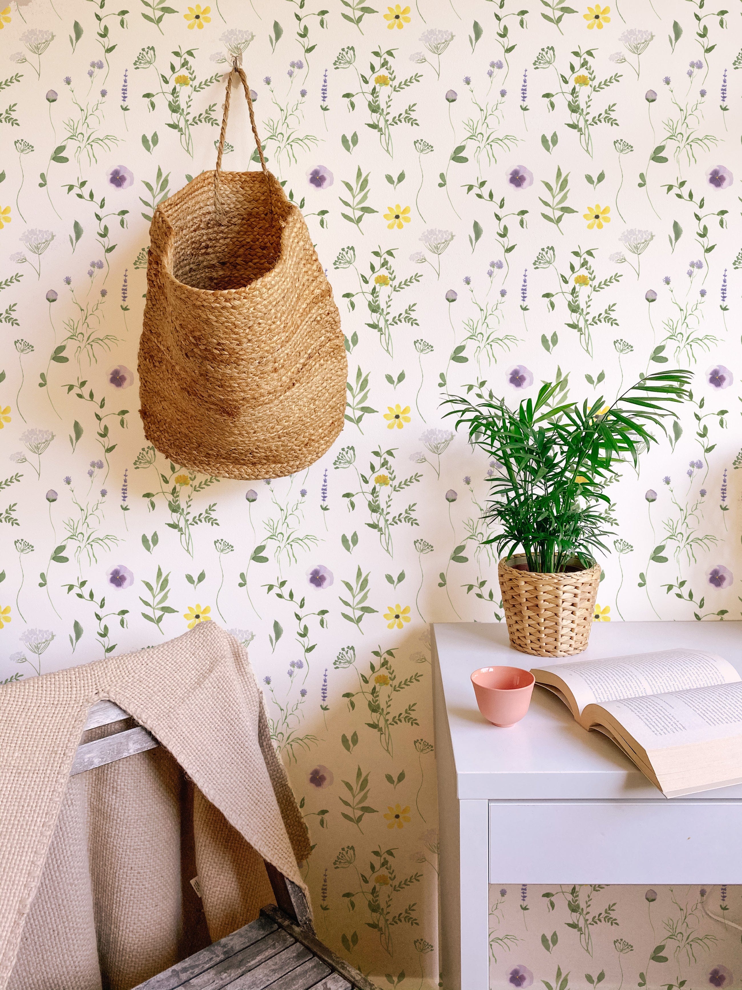 A cozy corner decorated with the Spring Field Wallpaper - VII, showcasing its charming pattern of purple and yellow flowers with green foliage. The space includes a woven basket hanging on the wall and a small potted plant on a white desk.