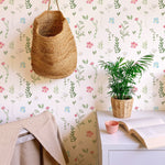 A cozy room featuring the Spring Field Wallpaper - VIII with a soft white background adorned with delicate pink, blue, and yellow flowers and green leaves. The decor includes a woven basket hanging on the wall and a small potted plant on a white desk