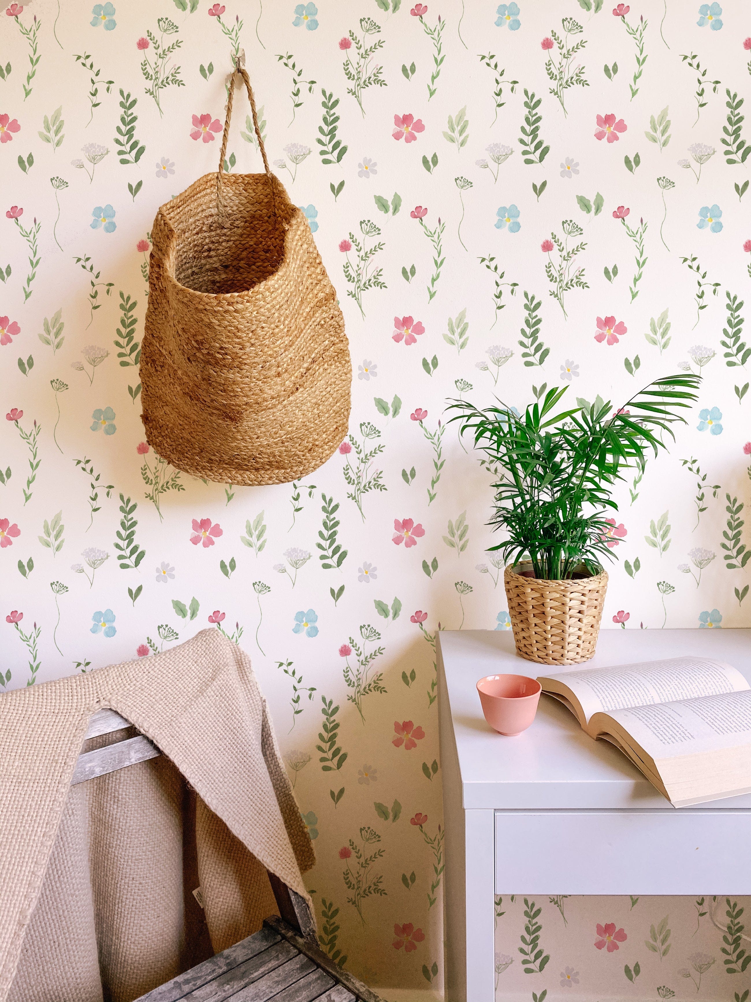 A cozy room featuring the Spring Field Wallpaper - VIII with a soft white background adorned with delicate pink, blue, and yellow flowers and green leaves. The decor includes a woven basket hanging on the wall and a small potted plant on a white desk