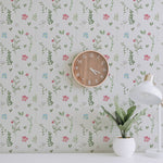 A workspace decorated with the Spring Field Wallpaper - VIII, showcasing its charming pattern of pink, blue, and yellow flowers with green foliage. The space includes a wooden wall clock and a white desk lamp beside a small plant in a white pot