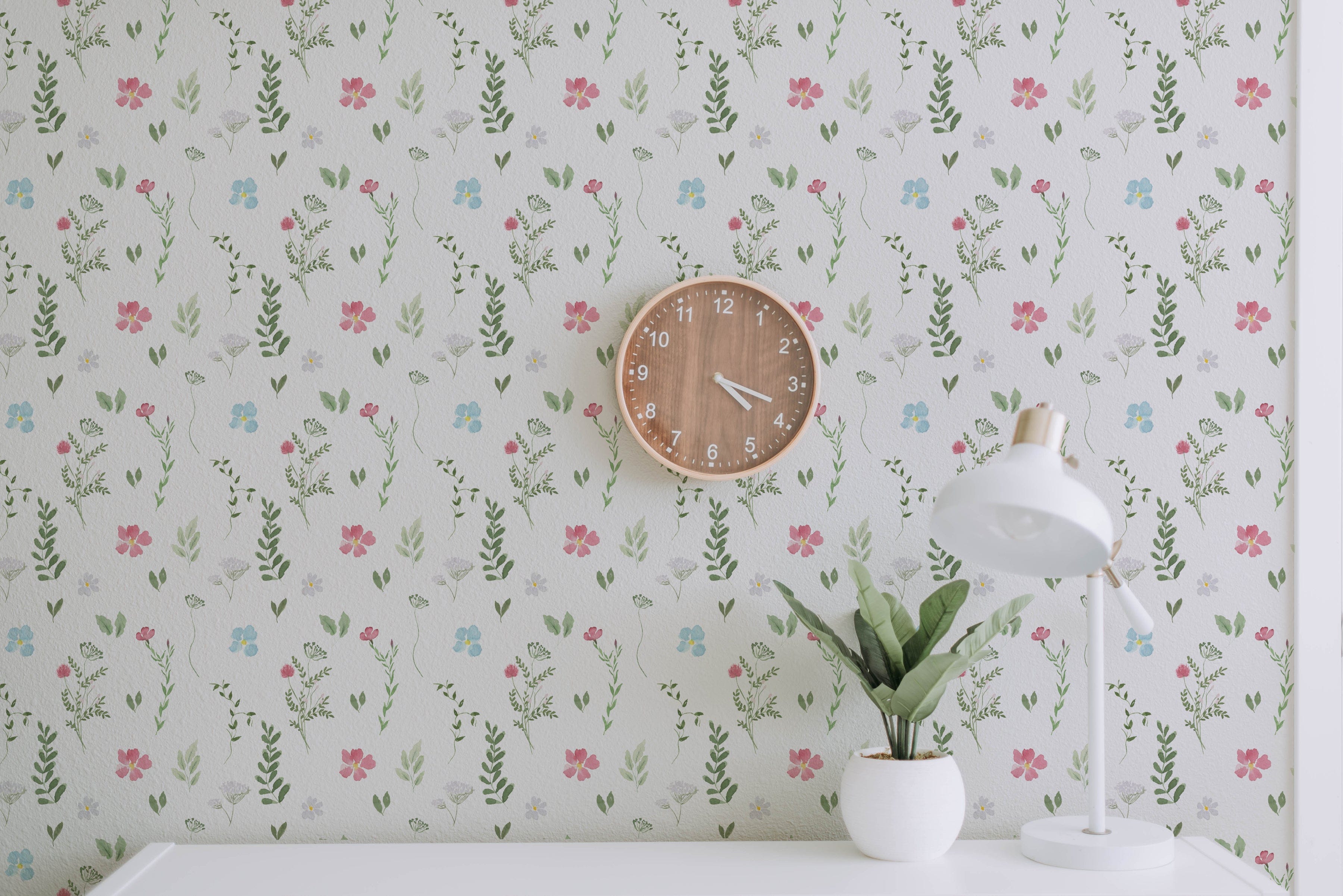 A workspace decorated with the Spring Field Wallpaper - VIII, showcasing its charming pattern of pink, blue, and yellow flowers with green foliage. The space includes a wooden wall clock and a white desk lamp beside a small plant in a white pot