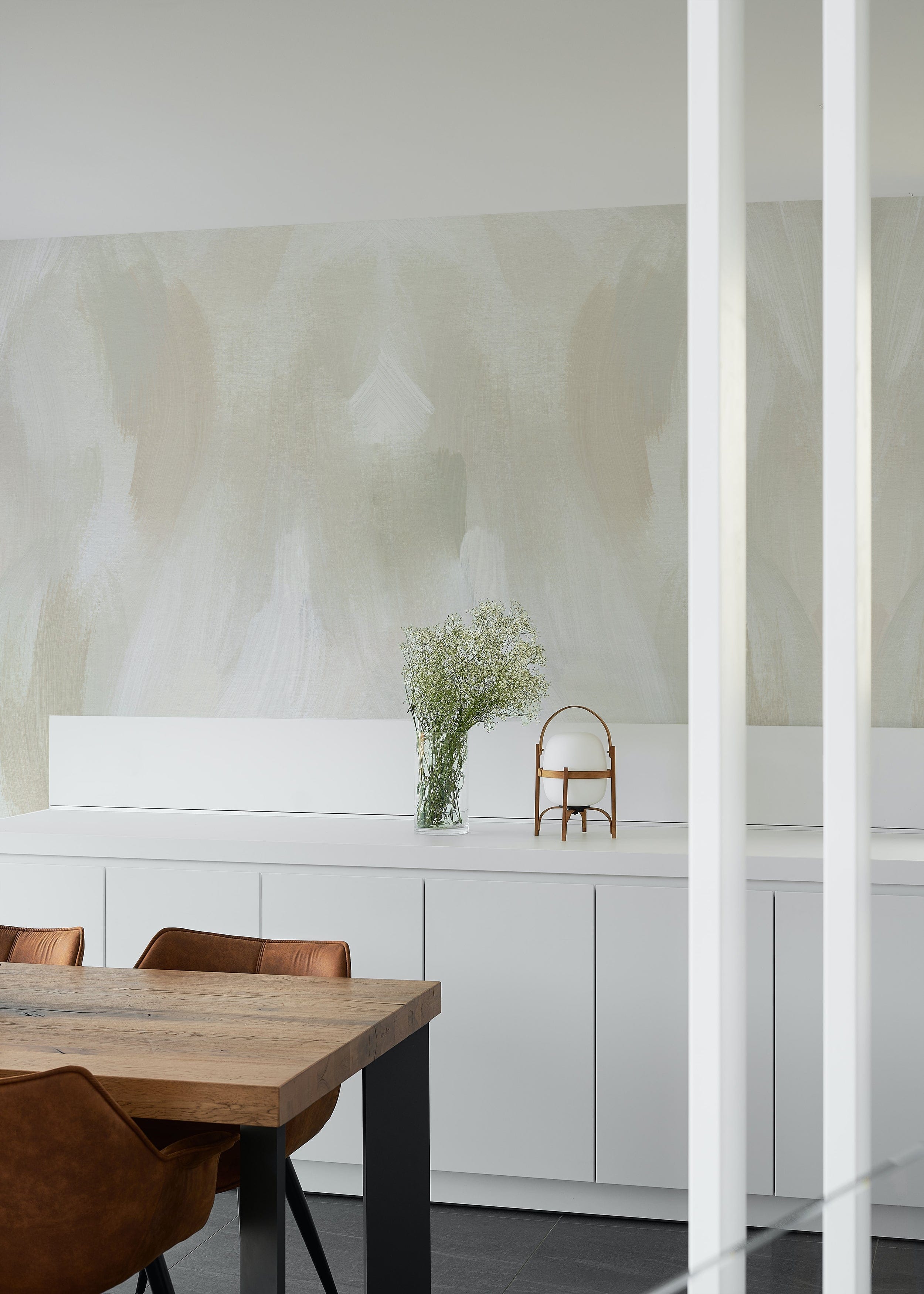 Earthy Aura Paint Texture Mural Wallpaper in a contemporary kitchen with a wooden dining table, leather chairs, white cabinets, and a vase of fresh flowers, highlighting the subtle and soothing paint texture design