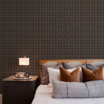 A luxurious bedroom accentuated by Dark Plaid Wallpaper - Black, providing a rich, textured backdrop to a bed with assorted pillows. The pattern adds depth and a traditional touch to the modern room, complemented by a sleek bedside table and a contemporary lamp.