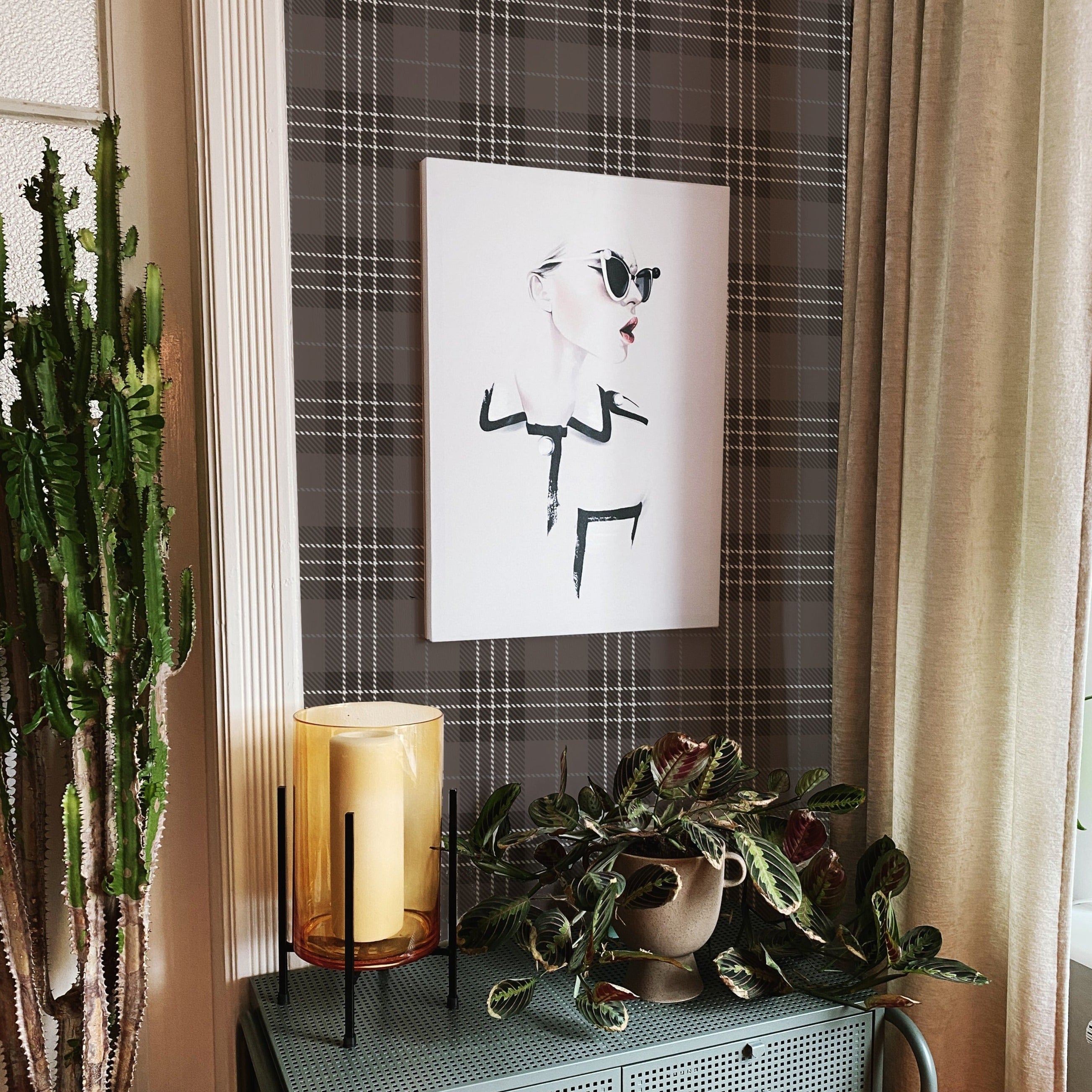 A snippet of home decor featuring the Dark Plaid Wallpaper - Black behind an art piece, flanked by indoor plants and a large candle, depicting an intimate corner that blends classic patterns with contemporary design elements for a unique, stylish ambiance.