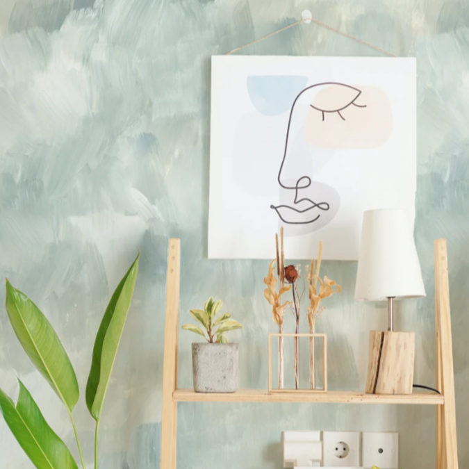 A living space with light blue and white textured wallpaper. The wallpaper has a cloud-like pattern with soft brush strokes. A wooden shelf stands against the wall, holding a framed abstract line drawing, a small plant, dried flowers, and a white lamp.
