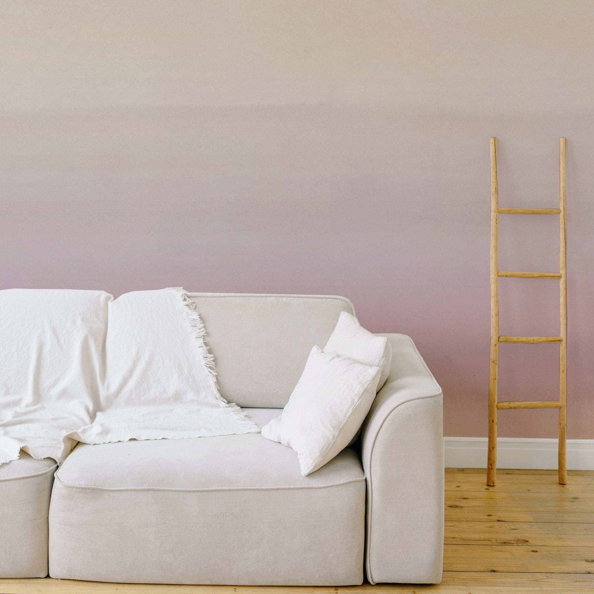 A cozy living room setting with a large, plush sofa against a wall adorned with Blush Gradient Wallpaper that fades from a creamy color to a gentle blush tone. A wooden ladder leans against the wall as a decorative element.