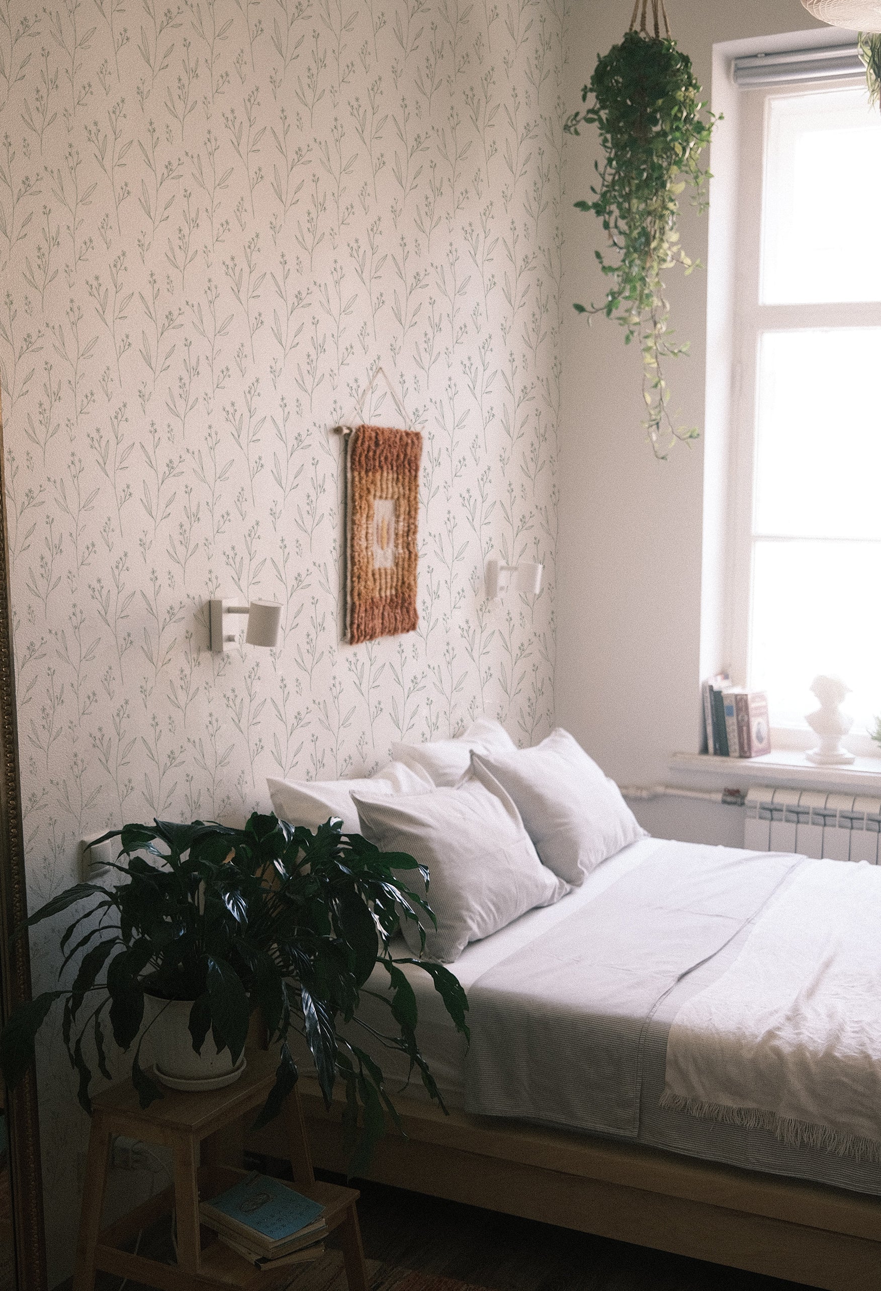 A light-filled bedroom corner with a minimalist aesthetic, featuring the Dainty Minimal Floral - Light Sage wallpaper. The wall exhibits a subtle and elegant pattern of light sage green floral illustrations against a clean, off-white background. A large, healthy potted plant sits by the white metal bed, which is neatly made with white linens, enhancing the room’s fresh and airy feel. A woven tapestry hangs above the bed, adding a touch of warmth and texture to the space.