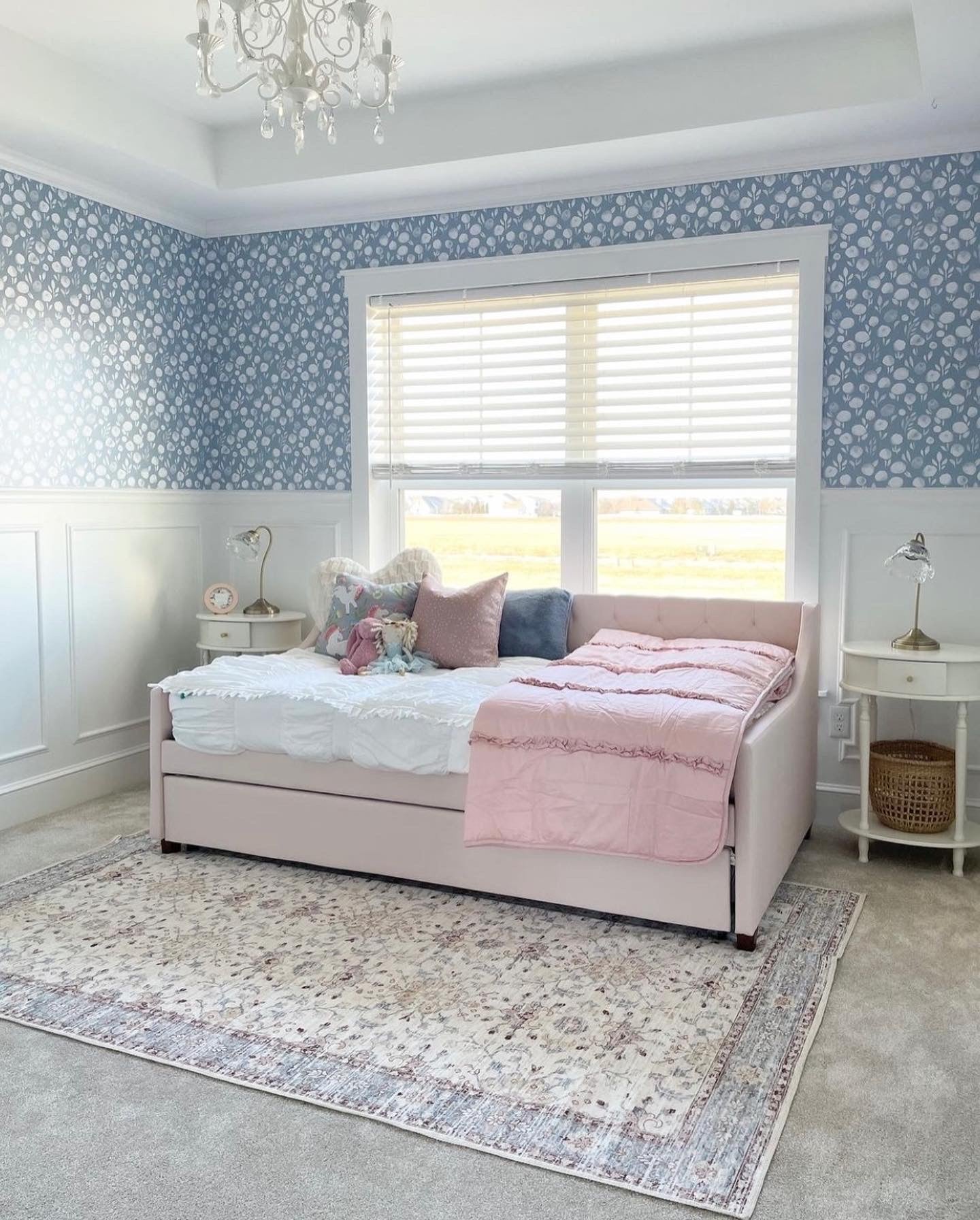 A light and airy bedroom with 'Subtle Botanica II - Inverted' wallpaper featuring a soothing pattern of white botanical silhouettes against a soft blue-grey background. The room is elegantly styled with a blush pink upholstered bed, a white bedside table, and a patterned rug, enhancing the serene aesthetic.
