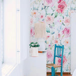 A bright, airy room featuring a wall adorned with vibrant Pink Floral Wallpaper. The floral design includes shades of pink, green, and white, complemented by a teal chair and a white dresser with a ceramic vase and lamp.