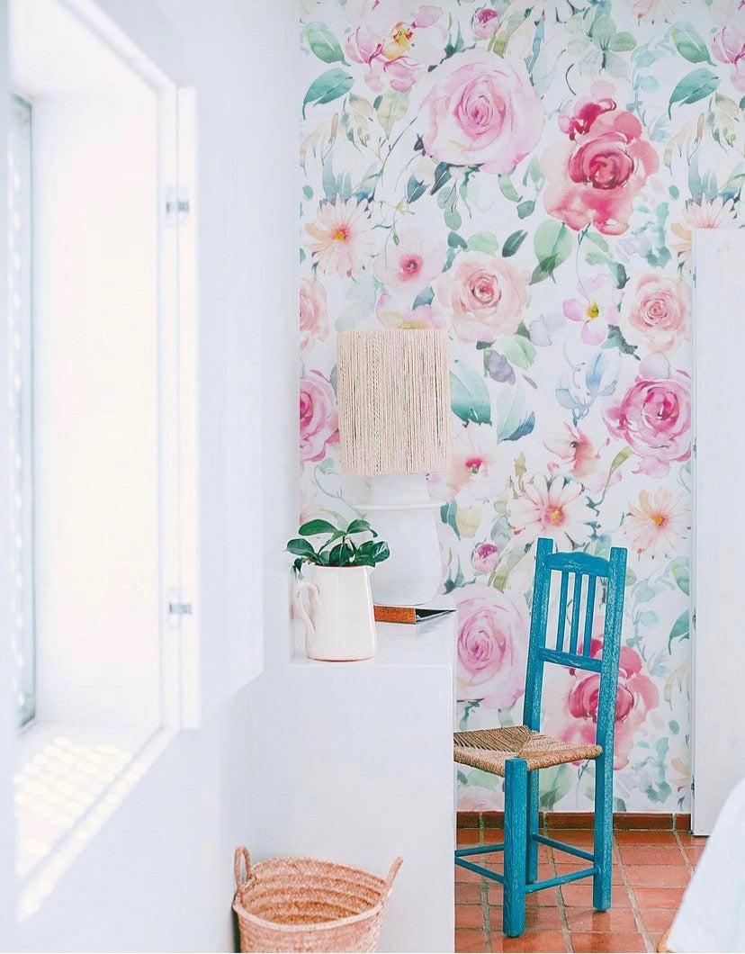 A bright, airy room featuring a wall adorned with vibrant Pink Floral Wallpaper. The floral design includes shades of pink, green, and white, complemented by a teal chair and a white dresser with a ceramic vase and lamp.