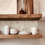 A close-up view of a kitchen shelf against a wall covered with Wildflower Sketch Wallpaper. The wallpaper displays an elegant black and white sketch of wildflowers, adding a touch of nature-inspired art to the space. On the wooden shelf, there is a neatly arranged set of white ceramic dishes including mugs, bowls, and plates. The rustic charm is complemented by the warmth of the wood and the clean lines of the tableware.