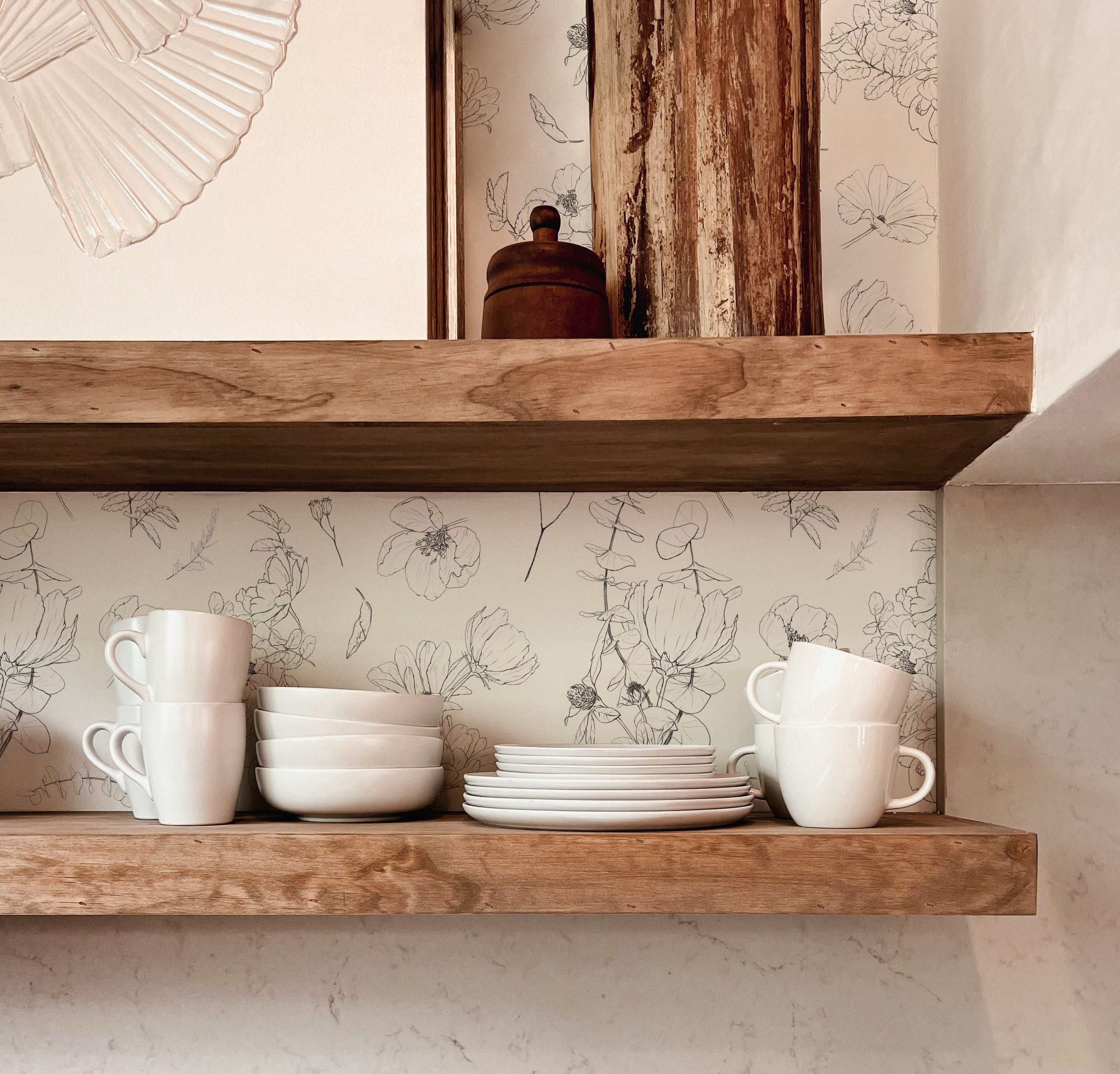 A close-up view of a kitchen shelf against a wall covered with Wildflower Sketch Wallpaper. The wallpaper displays an elegant black and white sketch of wildflowers, adding a touch of nature-inspired art to the space. On the wooden shelf, there is a neatly arranged set of white ceramic dishes including mugs, bowls, and plates. The rustic charm is complemented by the warmth of the wood and the clean lines of the tableware.